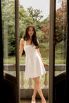 Woman in a white dress standing in front of glass doors with a garden view.