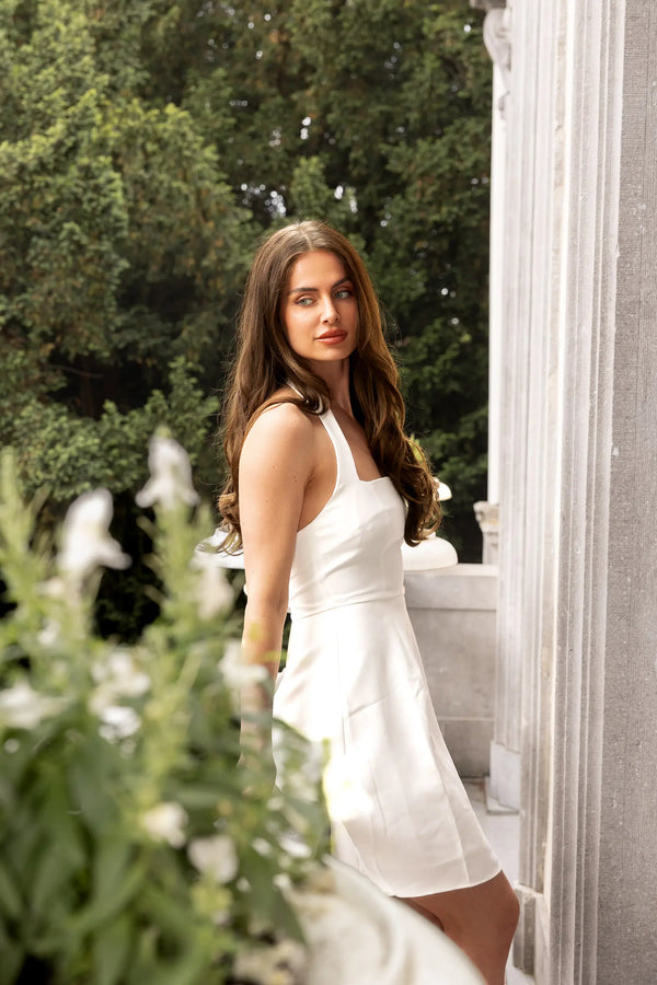 Woman in a white dress standing outdoors with greenery and architecture in the background