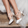 White high-heeled shoes with thin straps worn by a person in a white dress on stone steps.