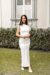 Woman in a white dress standing outdoors with greenery and a building in the background